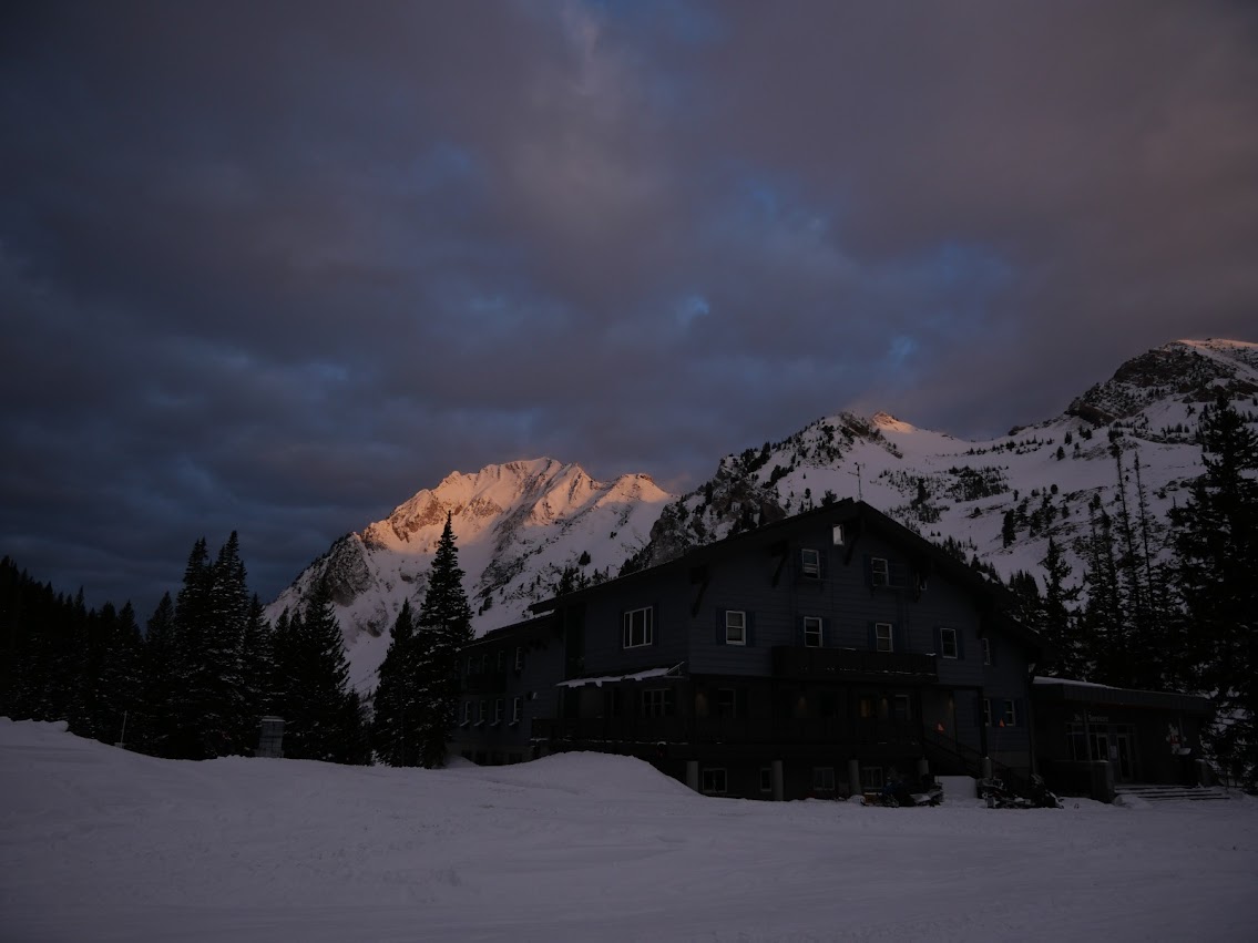 Alta Ski Lodge in the early morning. A ski lodge is the in foreground. In the background, a mountain peak is visible, with the first rays of morning sunlight on the peak.